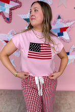 Load image into Gallery viewer, Woman wearing a pink shirt with an American flag design and red checkered pants against a colorful star-patterned wall.
