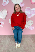 Load image into Gallery viewer, Woman wearing a red sweater and blue jeans standing in front of a pink wall with heart decorations.

