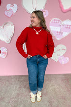Load image into Gallery viewer, Woman wearing a red sweater and blue jeans standing in front of a pink wall with heart decorations.
