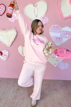 Load image into Gallery viewer, Person in pink sweatshirt with text, holding a bottle and teddy bear, against a pink wall with heart decorations.
