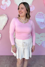Load image into Gallery viewer, Woman wearing a pink top and white skirt against a pink wall with heart decorations.

