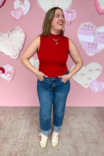 Load image into Gallery viewer, Woman in a red sleeveless top and blue jeans standing in front of a pink wall with heart decorations.
