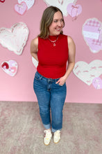 Load image into Gallery viewer, Woman wearing a red sleeveless top and blue jeans standing in front of a pink wall with heart decorations.
