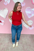 Load image into Gallery viewer, Woman wearing a red sleeveless top and blue jeans standing in front of a pink wall with heart decorations.
