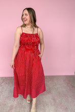 Load image into Gallery viewer, A woman modeling a red and white vertically striped midi dress with a smocked bodice, and beige strappy heels, set against a solid pink studio background.
