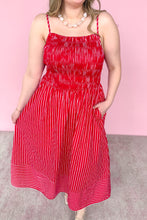 Load image into Gallery viewer, A woman modeling a red and white vertically striped midi dress with a smocked bodice, and beige strappy heels, set against a solid pink studio background.
