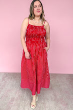 Load image into Gallery viewer, A woman modeling a red and white vertically striped midi dress with thin spaghetti straps, a smocked bodice, and side pockets, standing against a solid pink wall.
