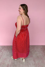Load image into Gallery viewer, A woman modeling a red and white vertically striped spaghetti-strap maxi dress, shown from a side-back angle against a solid pink wall.
