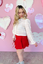Load image into Gallery viewer, Woman in a white blouse and red skirt standing against a pink wall with heart decorations side view.
