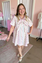 Load image into Gallery viewer, A woman wearing a short-sleeve pink and white floral print dress with a large bow at the collar, standing in a pink-themed room.
