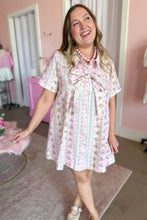 Load image into Gallery viewer, A woman wearing a short-sleeve pink and white floral print dress with a large bow at the collar, standing in a pink-themed room.
