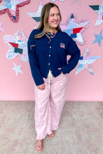Load image into Gallery viewer, Woman wearing a blue cardigan with an American flag design and pink pants against a pink wall with star decorations.
