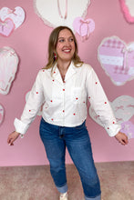 Load image into Gallery viewer, Woman wearing a white blouse with red hearts and blue jeans in front of a pink wall with heart decorations.
