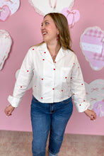 Load image into Gallery viewer, Woman wearing a white blouse with red hearts and blue jeans against a pink wall with heart decorations.
