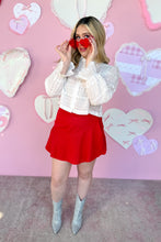 Load image into Gallery viewer, Woman in a white blouse and red skirt standing in front of a pink wall with heart decorations.
