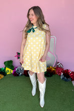 Load image into Gallery viewer, A woman modeling a yellow sleeveless dress with white polka dots and a green bow tie neckline, paired with white cowboy boots, standing on green artificial turf against a pink backdrop with holiday decorations.
